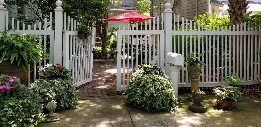 A white picket fence surrounds a private courtyard entry, featuring lush greenery and vibrant flowers. A red patio umbrella provides shade over a cozy seating area, inviting guests into a serene outdoor space that is carefully landscaped for privacy.