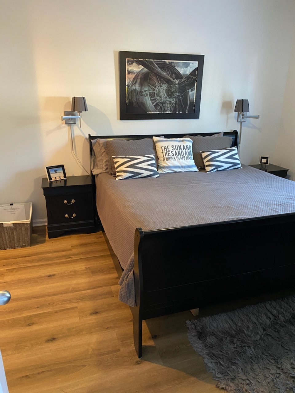 A well-furnished bedroom features a spacious bed with gray bedding and decorative pillows against a light wall. Two nightstands are positioned beside the bed, and wall-mounted lamps provide ambient lighting. A textured rug is placed on the warm wood floor, contributing to a cozy atmosphere.