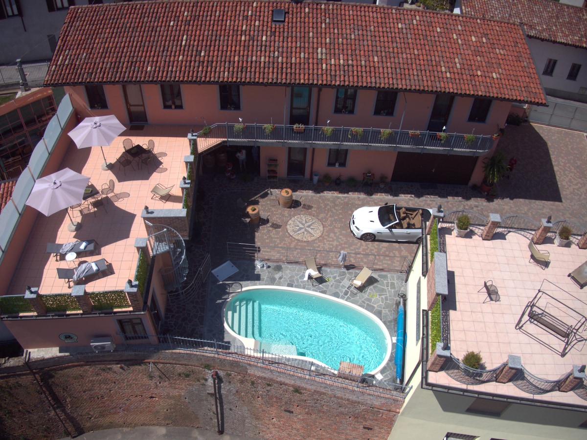 An aerial view of a charming property showcasing a uniquely shaped pool surrounded by a sun terrace. Sun loungers and umbrellas line the patio areas, while a classic car is parked nearby. The vibrant roof tiles and cobblestone pathways enhance the overall setting.