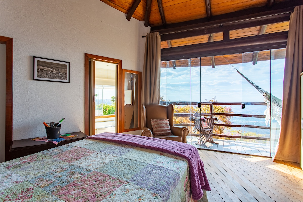 A bright bedroom features a spacious bed covered with a patterned quilt, alongside a comfortable armchair. Large glass doors open to a balcony with a hammock, offering expansive views of the natural surroundings. Wooden flooring complements the warm wooden ceiling.