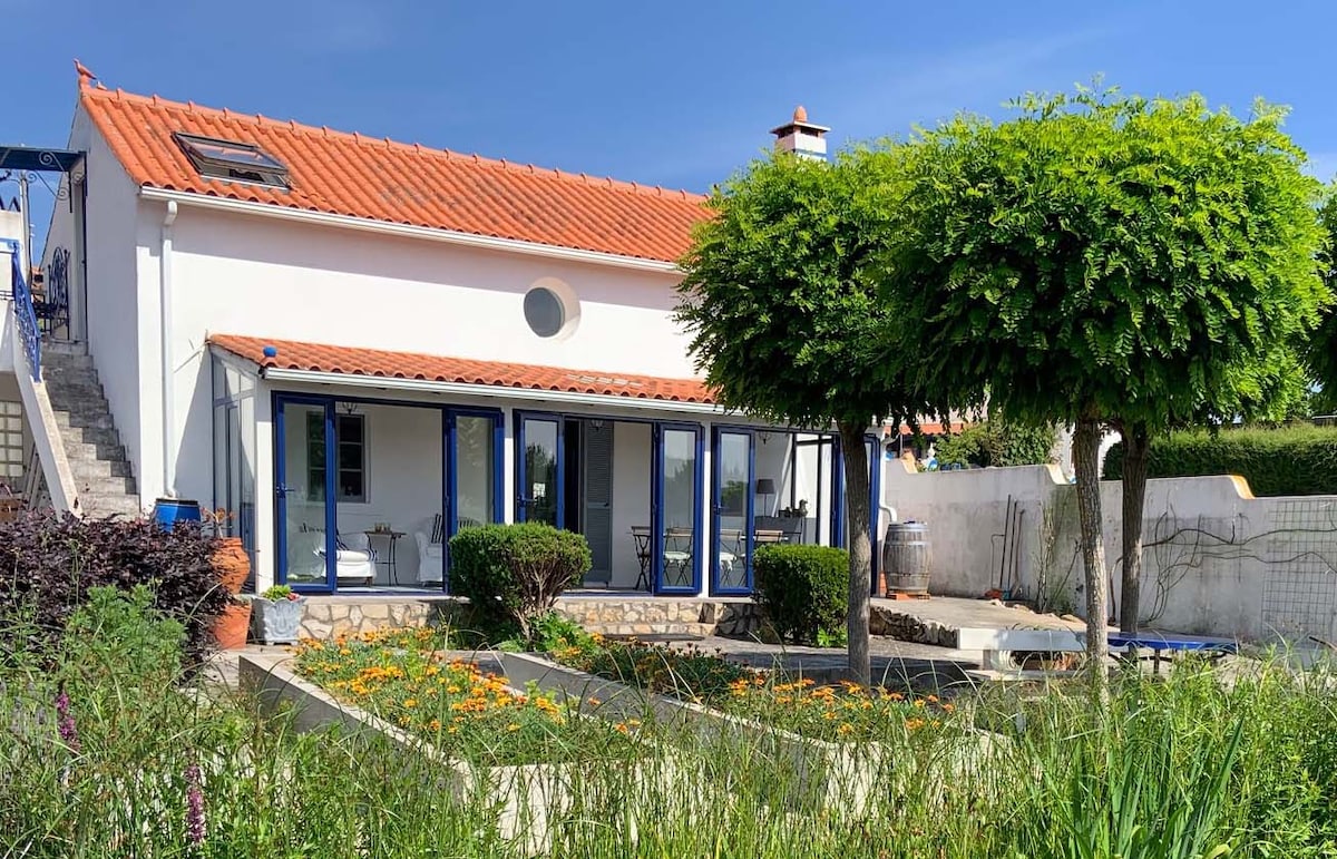 A charming cottage features a terracotta roof and large glass doors opening to a landscaped garden. Bright flowers and greenery surround the outdoor area, creating a serene environment. A staircase leads to the entrance, and a tree offers shade on a sunny day.
