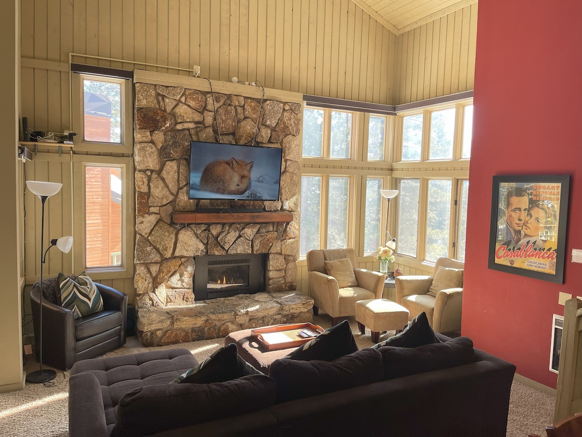 A spacious living area features a stone fireplace surrounded by large windows that allow ample natural light. Comfortable seating includes a dark sofa, two light-colored armchairs, and a coffee table. A wall-mounted TV is positioned above the fireplace, enhancing the cozy ambiance.