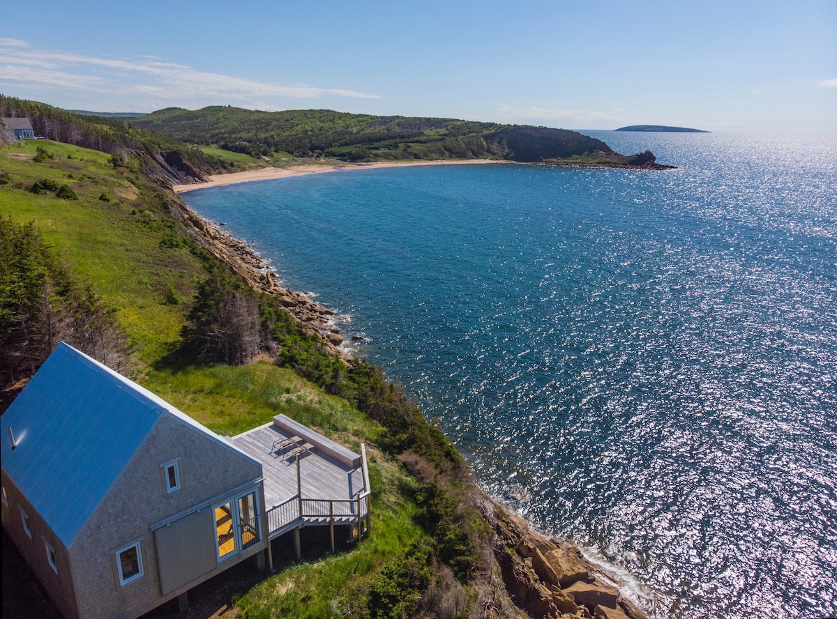 An aerial view of the charming cottage perched on a cliff overlooking the ocean, with a spacious deck extending toward the water. The coastline and sandy beach stretch out in the distance, framed by lush green hills under a clear blue sky.