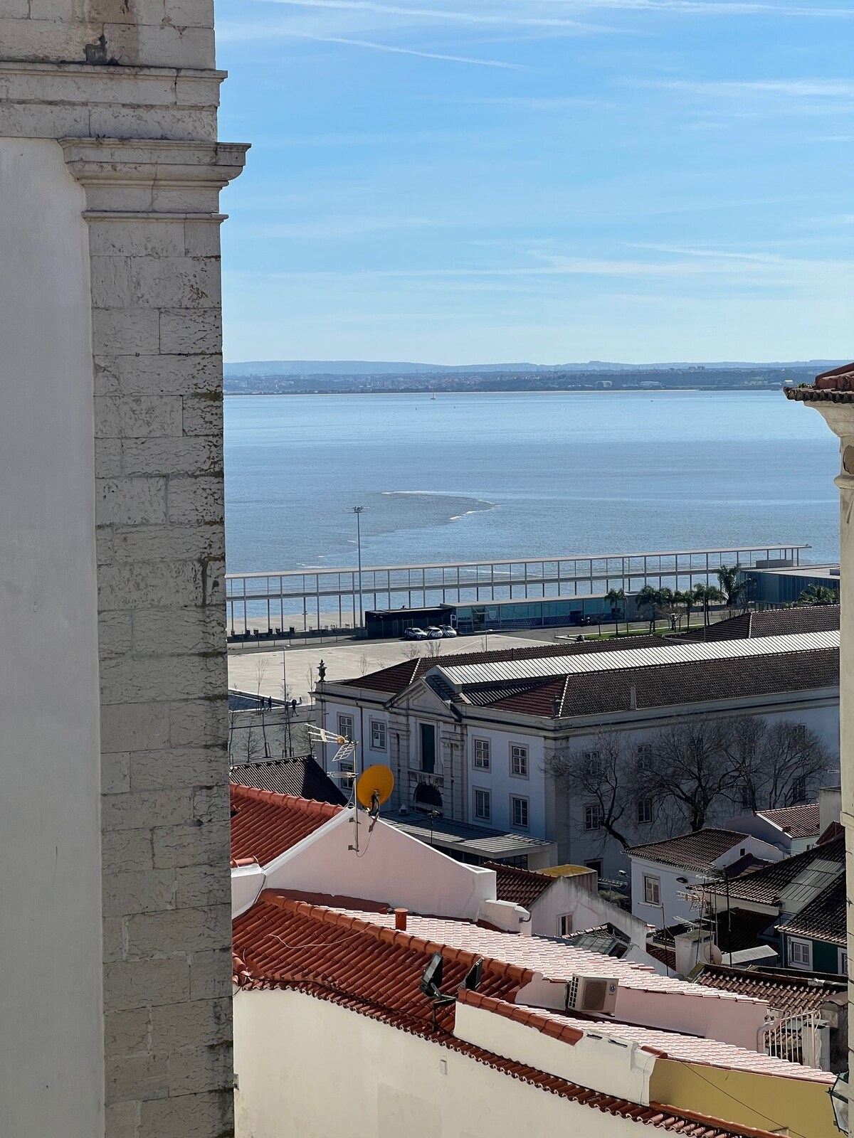 A sweeping view of the river Tejo is captured from an elevated position, showcasing the water's surface glistening under sunlight. Traditional red-tiled roofs of nearby buildings are visible in the foreground, with a glimpse of the waterfront and distant landscape beyond.