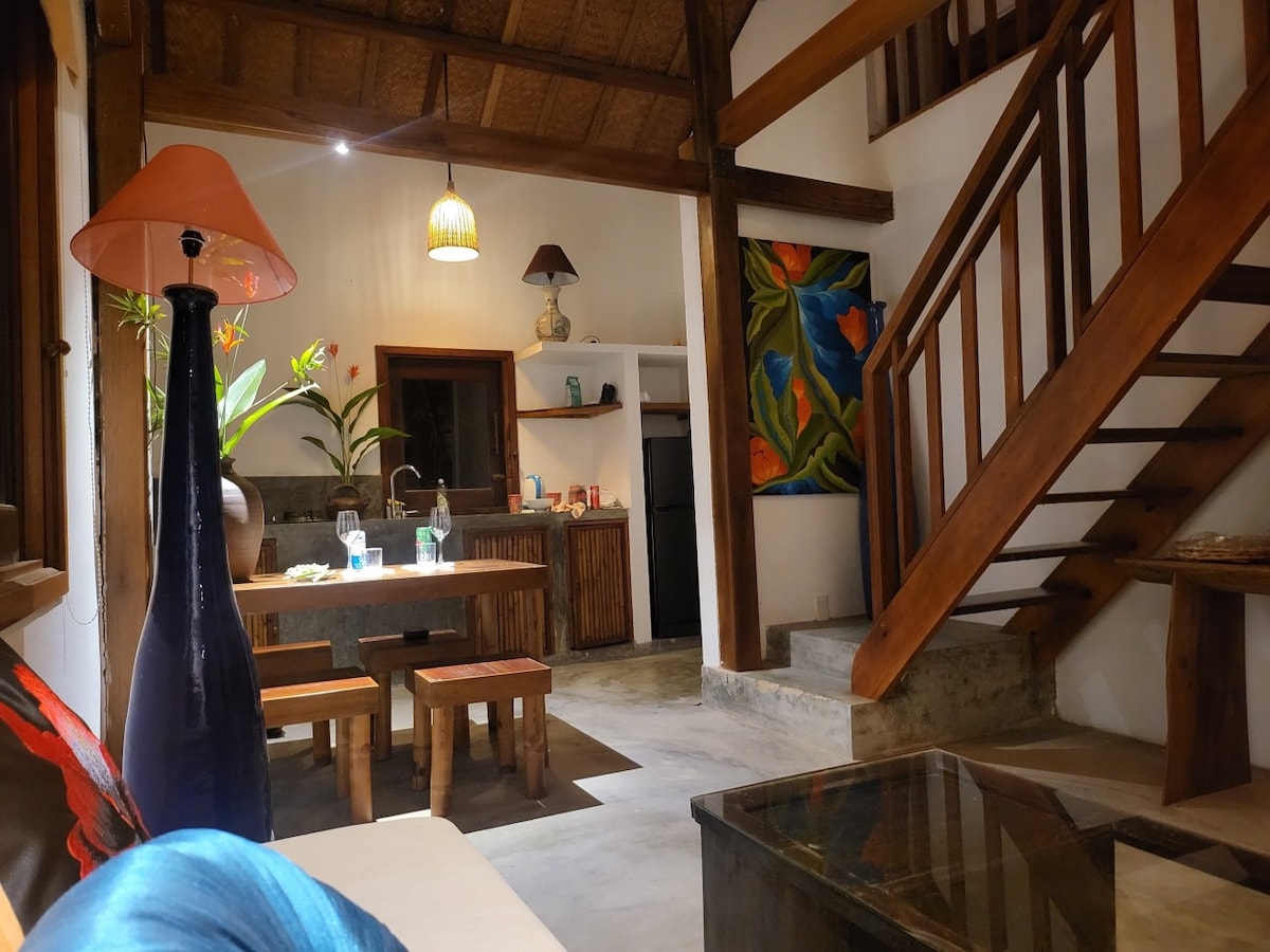 An inviting open-plan living area features wooden beams and a staircase leading to the upper floor. A dining table is positioned near the kitchen, which includes modern appliances. Decorative plants complement the space, with artistic touches visible on the walls.