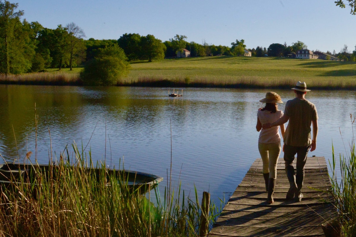 A serene dock extends over a calm body of water, bordered by lush greenery and tall grasses. Two individuals are seen walking side by side, enjoying the tranquil setting under clear blue skies. In the distance, a small rowboat is visible on the water.