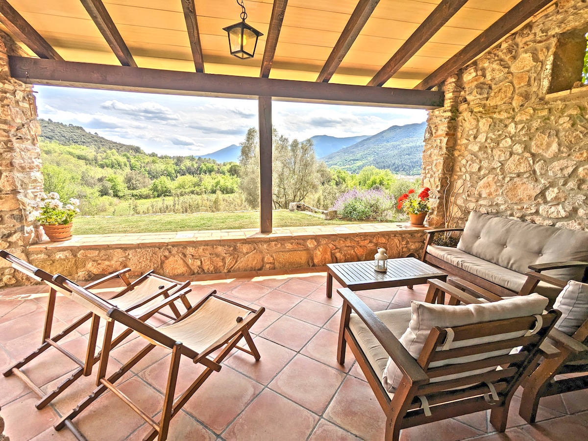 A covered outdoor seating area features wooden furniture, including a sofa and two reclining chairs. Potted plants add a touch of color, while expansive windows offer views of rolling hills and lush greenery. The stone walls and warm tile floor create a cozy atmosphere.