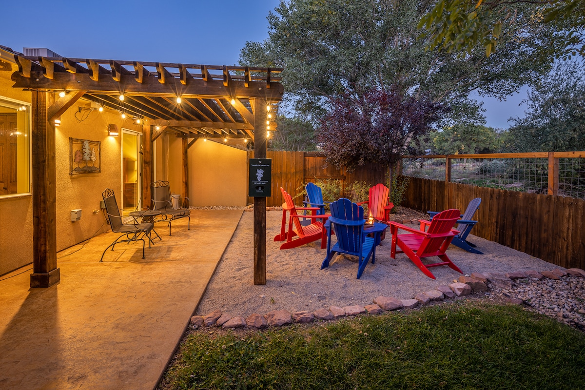 A cozy outdoor seating area features a pergola adorned with string lights. Brightly colored adirondack chairs in red and blue surround a firepit on a sandy patio, creating an inviting space for gathering. A lush green lawn and tall trees frame the area.