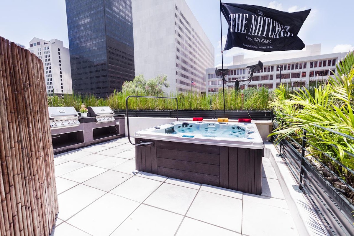 An outdoor area featuring a hot tub surrounded by a tiled patio. Gas BBQ grills are positioned nearby, offering a convenient space for grilling. Lush greenery lines the area, enhancing the natural ambiance, while a flag bearing the property name adds a touch of character.