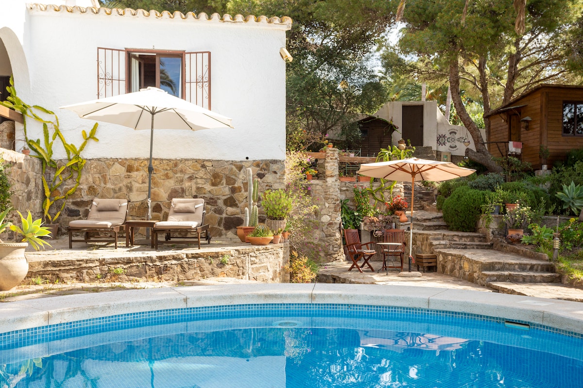 A tranquil pool area is framed by natural stone walls and lush greenery. Two lounge chairs and a small table are positioned under umbrellas. Stone steps lead to a cozy garden space with additional seating, creating an inviting outdoor retreat.