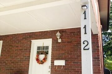 The entryway features a brick facade with a white front door adorned with a seasonal wreath. A charming light fixture is mounted above the door, while house number '112' is displayed prominently on the column beside it.