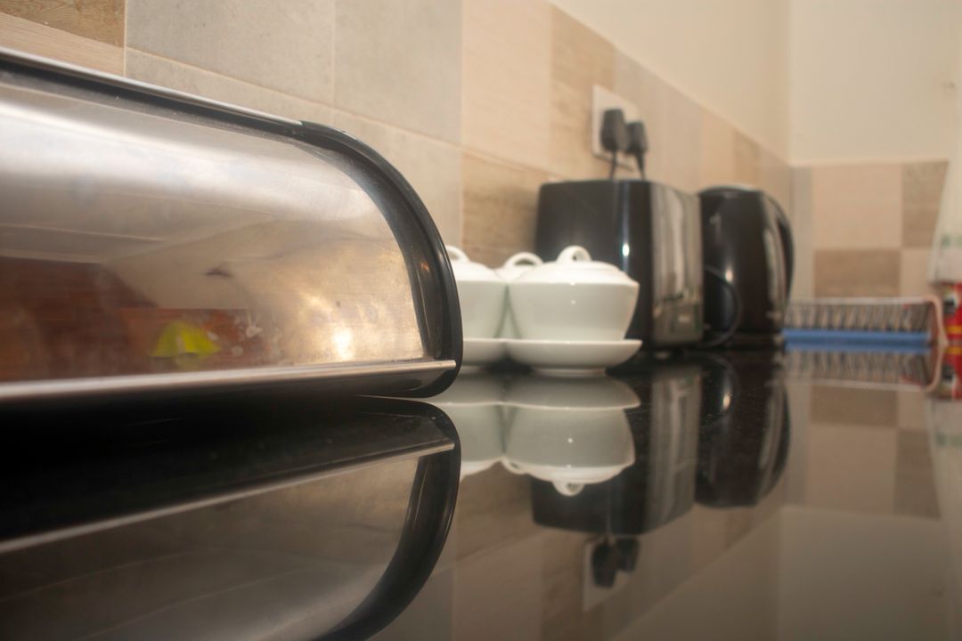 A sleek kitchen counter is displayed, showcasing a variety of appliances including a silver toaster, black kettles, and neatly arranged white dishes. The smooth black surface reflects the elements above, contributing to a clean and organized appearance.