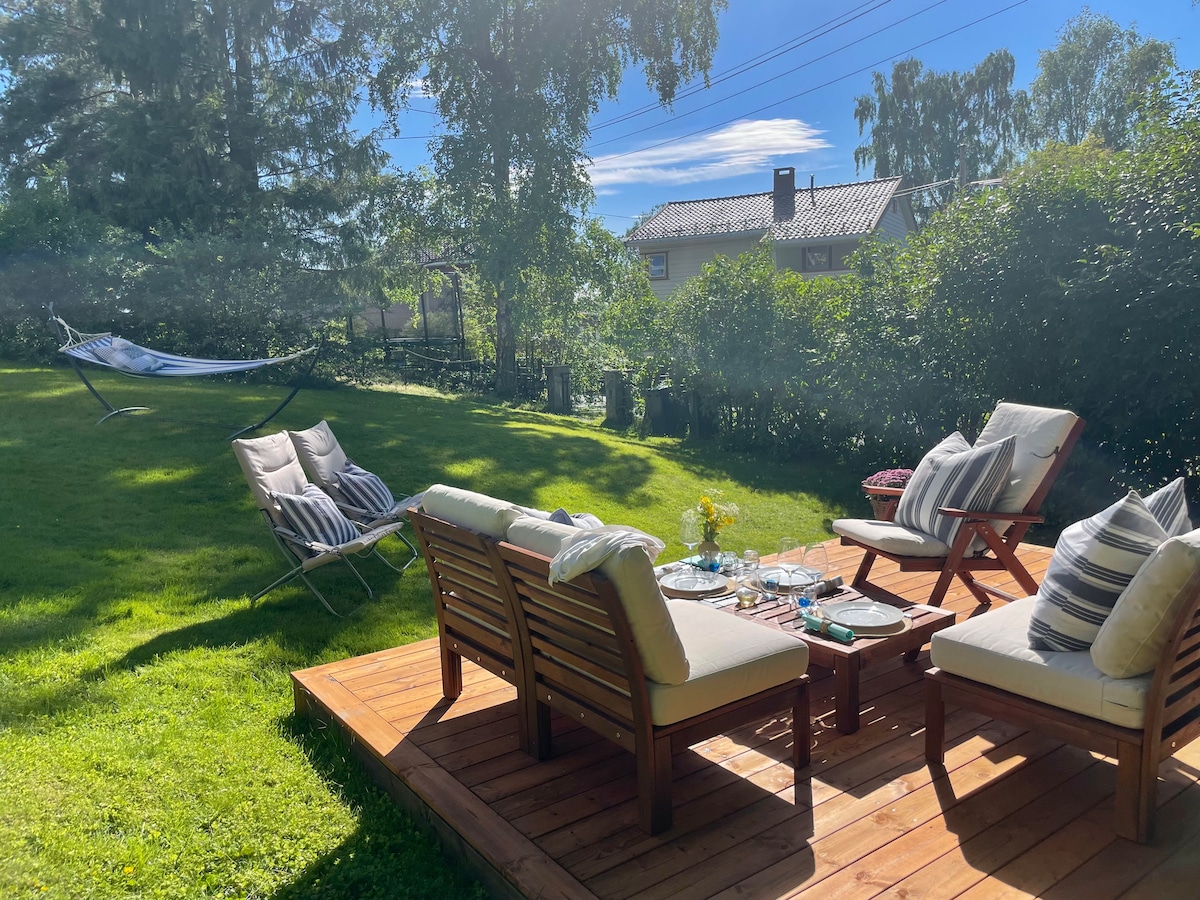 A wooden deck is surrounded by greenery, featuring a seating area with cushioned chairs and a table set with tableware. In the background, a hammock is visible among the trees, offering a relaxed spot in the sunlit garden.