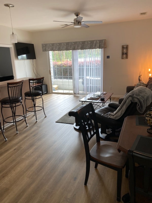 A living area is shown featuring a comfortable couch and a large window with sheer curtains allowing natural light. A dining table with black chairs and barstools at a counter are visible. Wood flooring adds warmth to the space.