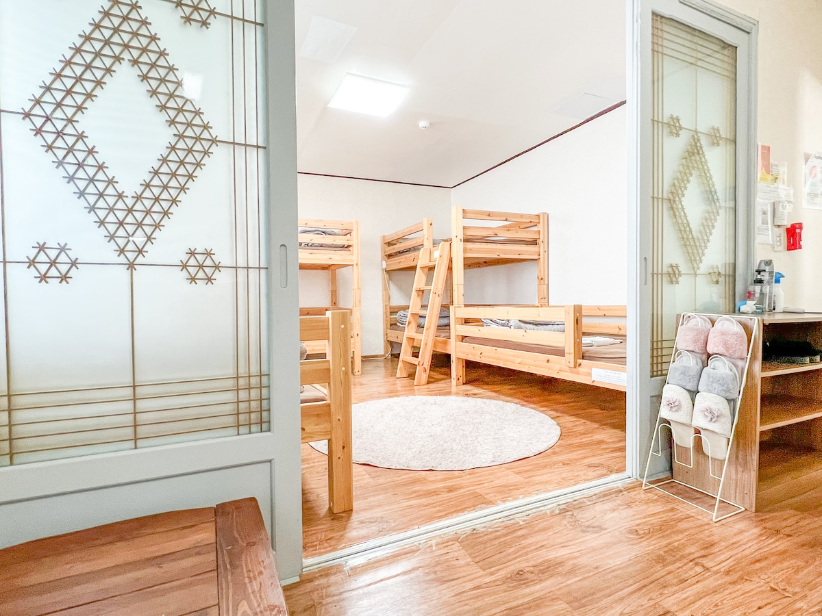 A spacious room is revealed through frosted glass doors, featuring wooden bunk beds arranged thoughtfully. A circular area rug sits in the center, with natural wood flooring enhancing the bright atmosphere. A shelf displays neatly stacked towels, adding to the inviting comfort of the space.