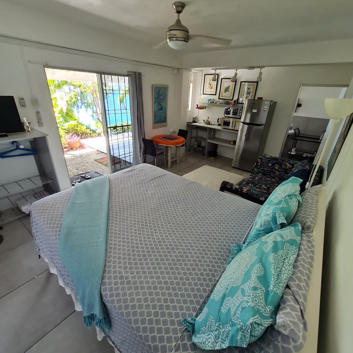 The studio apartment features a spacious bed with decorative pillows, complemented by a couch and a kitchenette area. Natural light enters through glass doors leading to the outside patio, where views of the garden are visible. An inviting atmosphere is created by the light-colored walls.