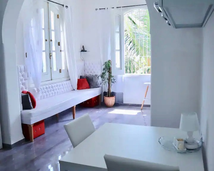 A bright living area features a white tufted bench and a small dining table. Large windows allow natural light to fill the space, framed by white curtains. A potted plant adds a touch of greenery, contributing to the airy atmosphere.