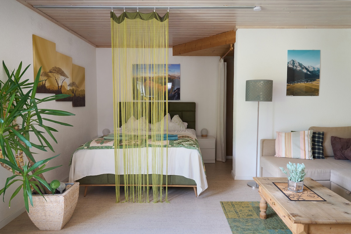 A spacious sleeping area is divided by a vibrant yellow curtain, creating a sense of privacy. A double bed is adorned with a decorative throw. Natural light floods the room, highlighting the green accents and inviting seating area with a coffee table.