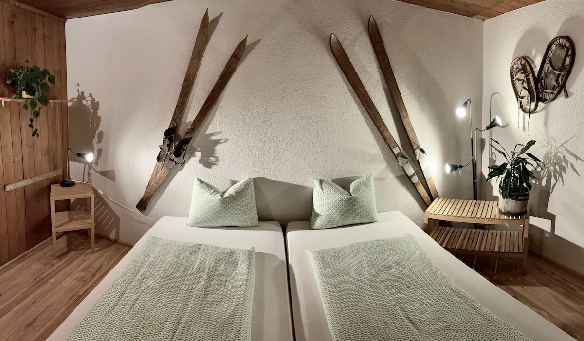 The sleeping area features two spacious beds with mint green bedding, positioned side by side. Wooden accents and ski equipment hang gracefully on the wall. A wooden side table holds a plant, while modern lamps provide soft lighting, enhancing the space's cozy atmosphere.