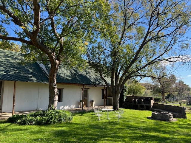 A serene outdoor area features a spacious lawn shaded by a large tree. A cozy seating arrangement with white chairs is positioned in front of the cottage, which has a simple façade and a green roof. Natural greenery surrounds the peaceful retreat.