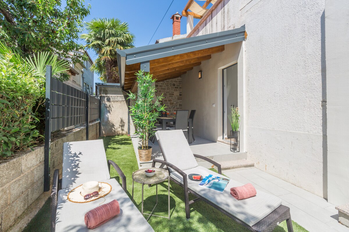 An outdoor area features two lounge chairs paired with a small table, positioned on well-maintained grass. A covered patio provides shade, showcasing a dining set, with greenery surrounding the space, including potted plants and trees.