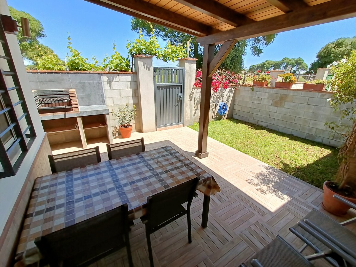 A private garden area is enclosed by walls adorned with greenery. A shaded seating space features a patterned table and four chairs. A barbecue is positioned in one corner, while vibrant flowers add color. The space offers a serene outdoor setting for relaxation.