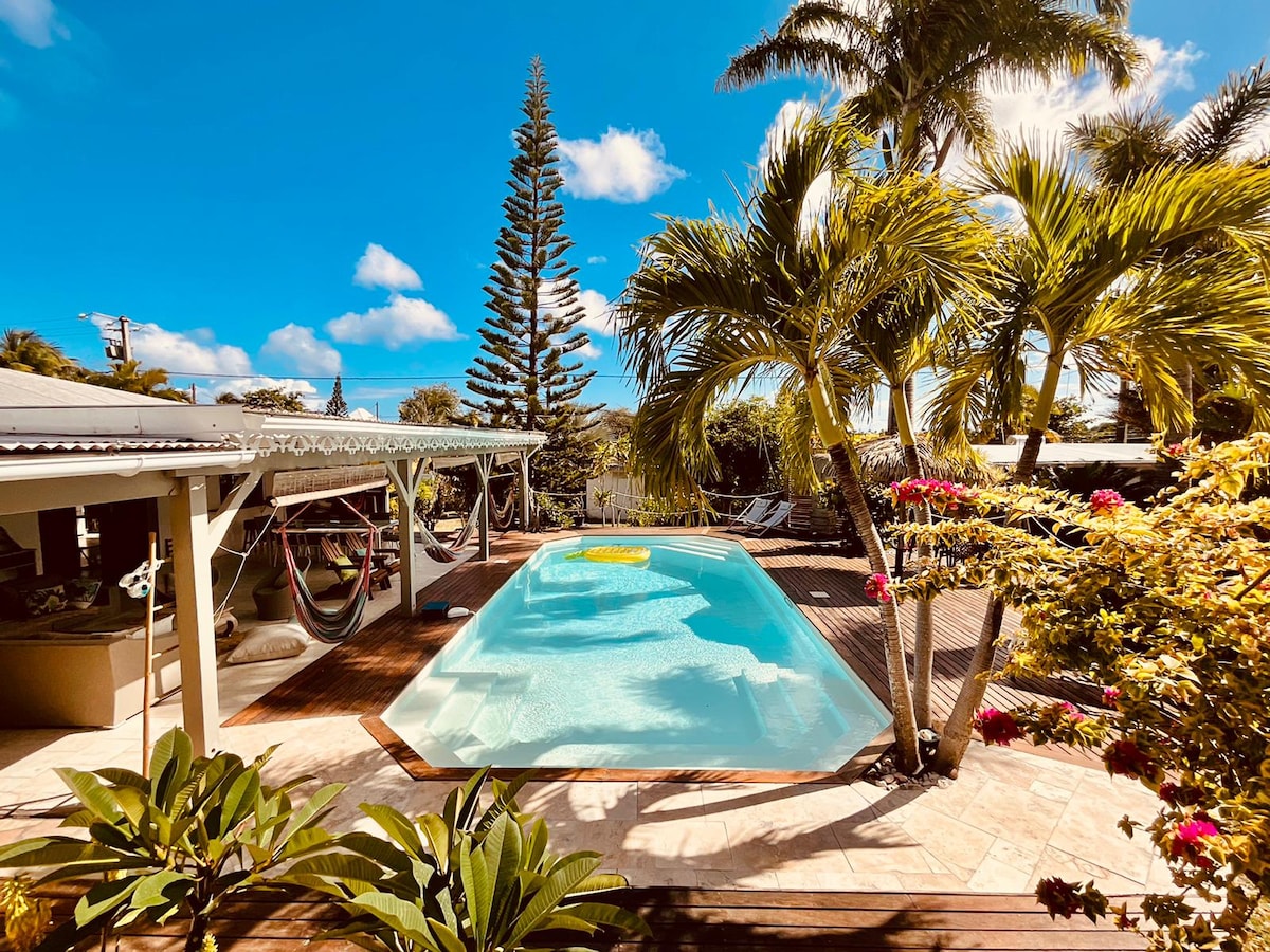 A bright outdoor space features a salt-water pool surrounded by tropical plants and palm trees. Comfortable lounge chairs and hammocks are visible, inviting relaxation. The sun illuminates the area, highlighting the deck's natural wood tones and inviting ambiance.