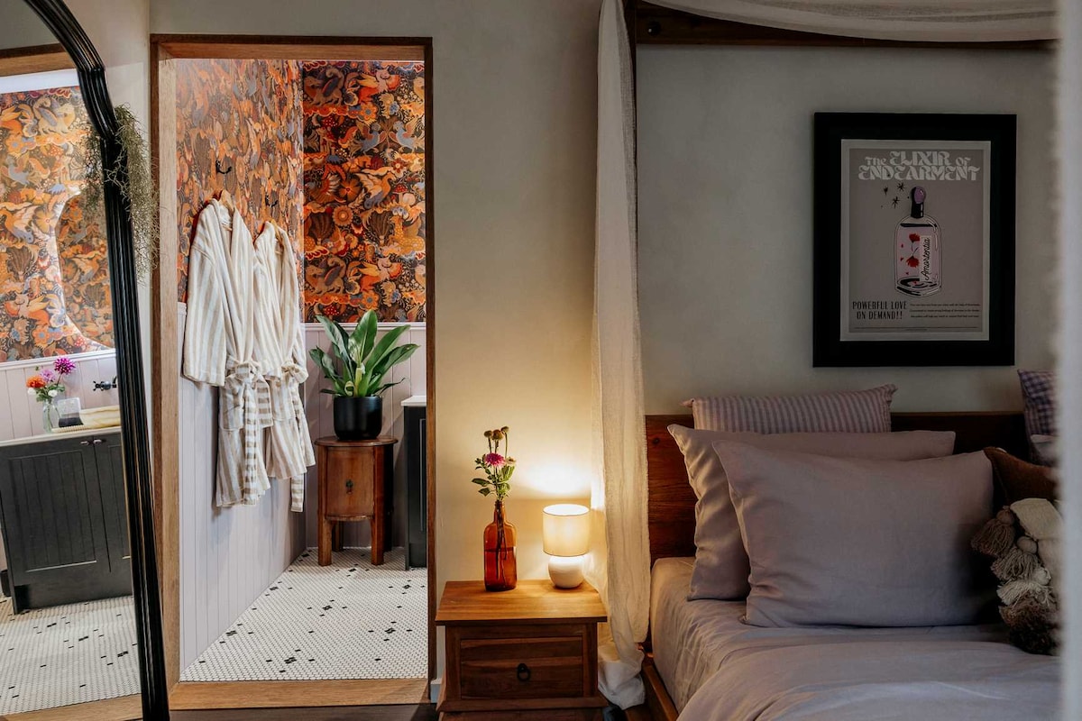 A cozy bedroom setting is visible, featuring a spacious bed adorned with soft linens. A bedside table holds a lamp and a floral arrangement. In the background, a doorway leads to a bathroom, accented by warm tones and decorative wall art.