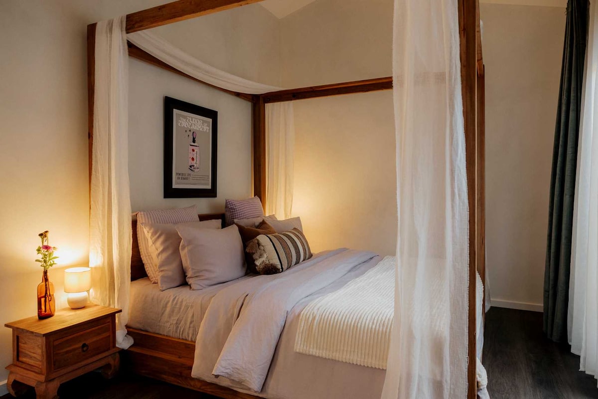 A queen-sized bed, draped with sheer curtains, is positioned against a softly lit wall. A wooden bedside table holds a small vase with a flower, while a thoughtfully arranged selection of pillows in neutral tones enhances the inviting appearance of the bedding. Light filters through the nearby window.