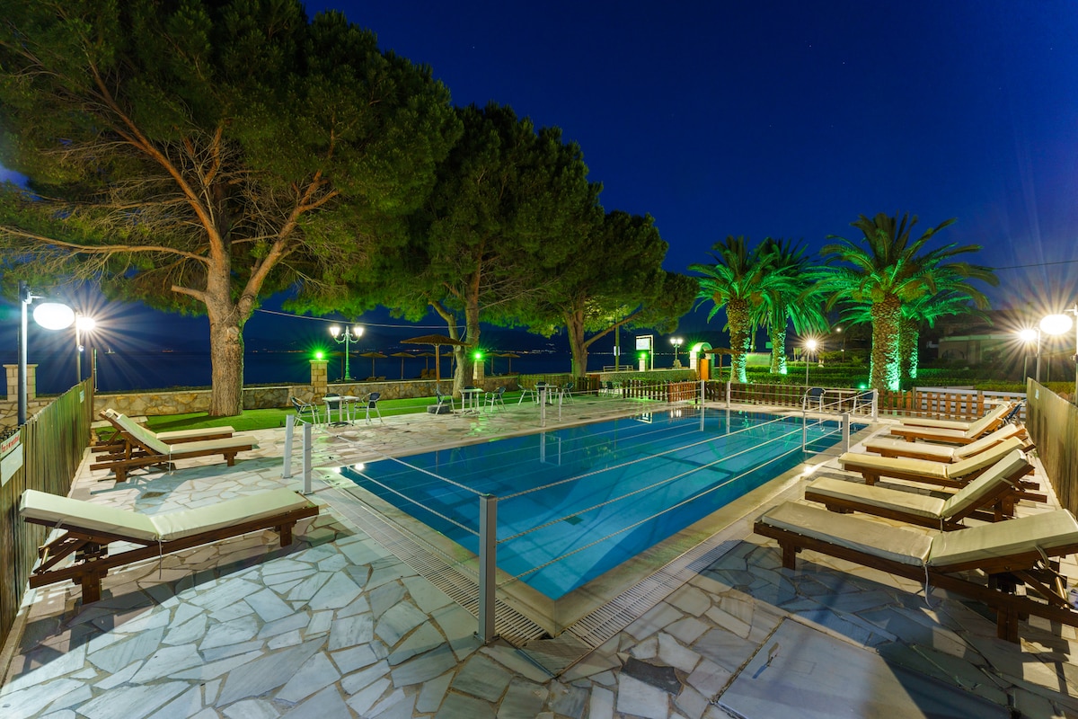 An outdoor swimming pool is surrounded by stone pathways, lounge chairs, and palm trees. Soft lighting enhances the setting, creating a serene ambiance. The pool area is spacious and well-maintained, with steps leading into the inviting water.