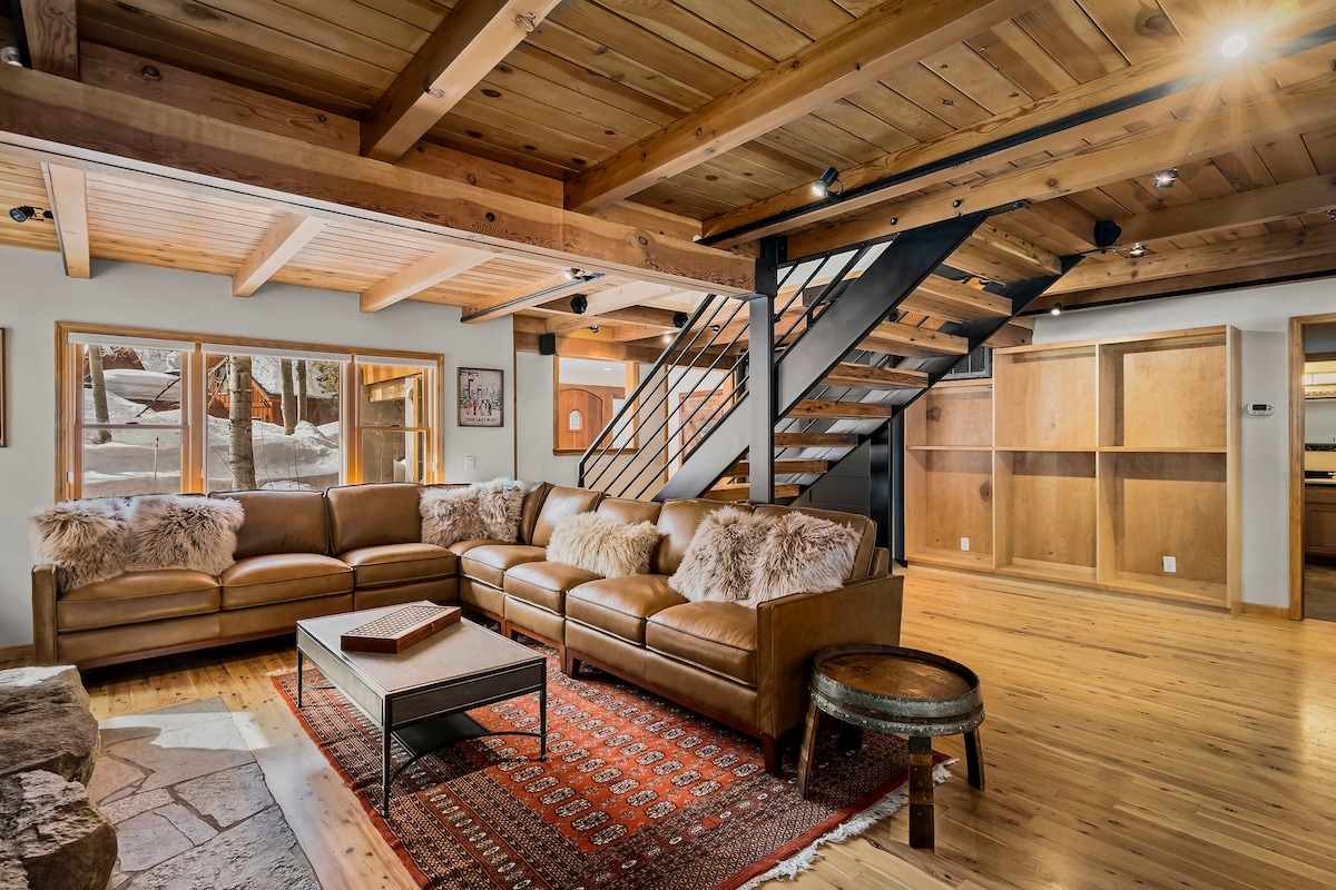 A spacious living area is highlighted by a leather sectional couch featuring decorative pillows. A wooden coffee table sits atop a patterned area rug. Large windows provide natural light, framing views of the outdoors. The ceiling beams and wooden flooring add warmth to the space.