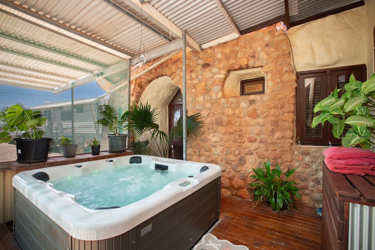 An enclosed spa area features a modern hot tub surrounded by greenery and decorative plants, creating a serene environment. Natural stone walls provide a rustic backdrop, while a shaded roof offers protection from the elements.