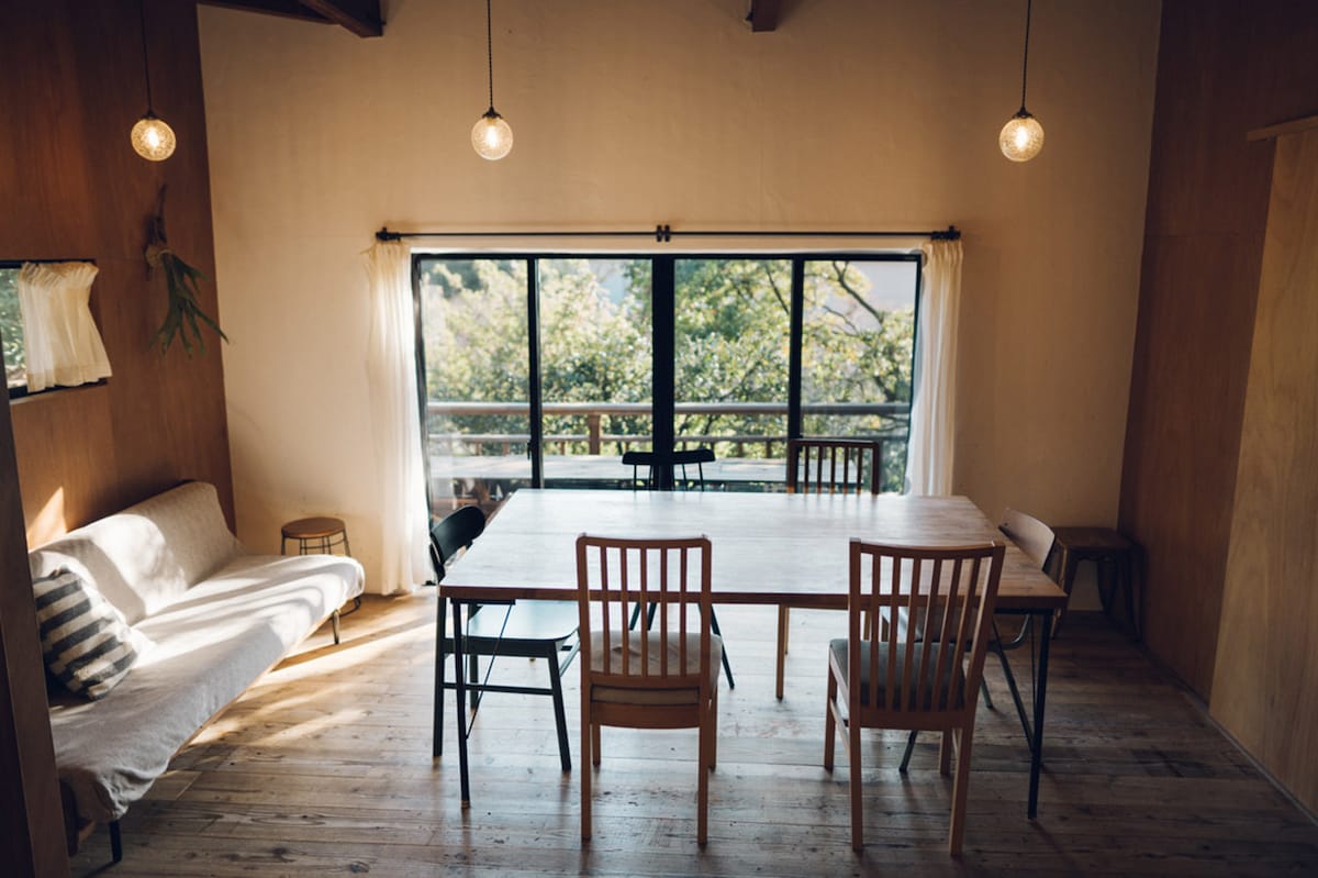 A spacious dining area features a large wooden table surrounded by several chairs. Natural light streams through large glass doors, illuminating the room and providing a view of the outside greenery. A cozy sofa is placed against the wall, adding to the inviting ambiance.