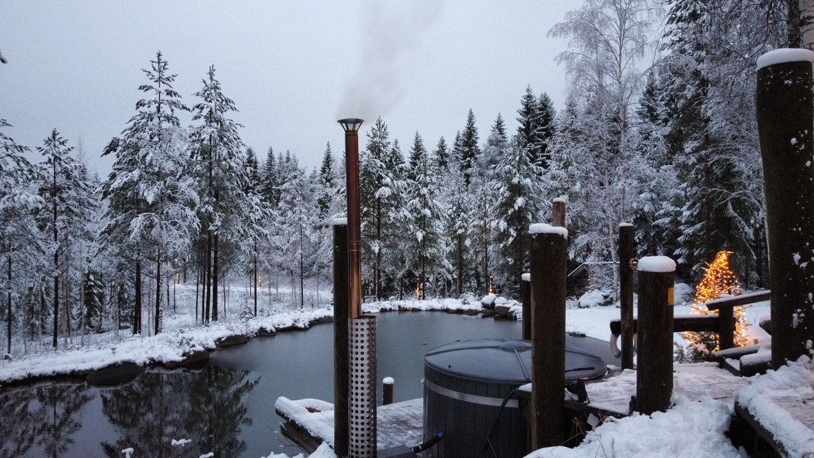 A serene winter landscape features a hot tub beside a gently steaming outdoor sauna. Snow-covered trees surround a still pond, creating a tranquil setting. Soft lights from nearby structures add a warm contrast to the cool hues of the snowy scene.