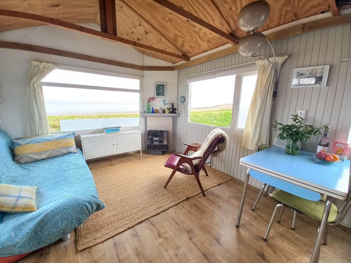 A bright living area is highlighted by large windows offering views of the surrounding landscape. A comfortable sofa with a light blue cover is positioned beside a small table set with fresh fruit. A vintage armchair and a compact fireplace complete the cozy ambiance.
