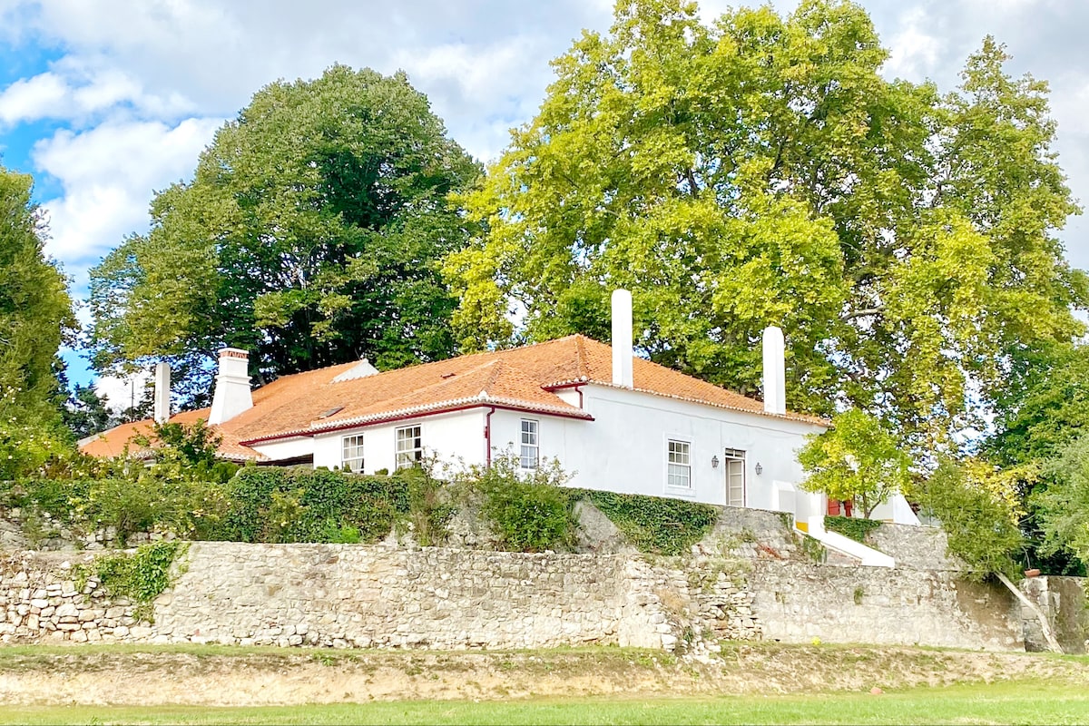 A two-story white house with a red-tiled roof stands prominently in a lush green landscape. Large trees surround the property, providing shade and a sense of tranquility. The structure features multiple chimneys and several windows, blending harmoniously with the natural surroundings.