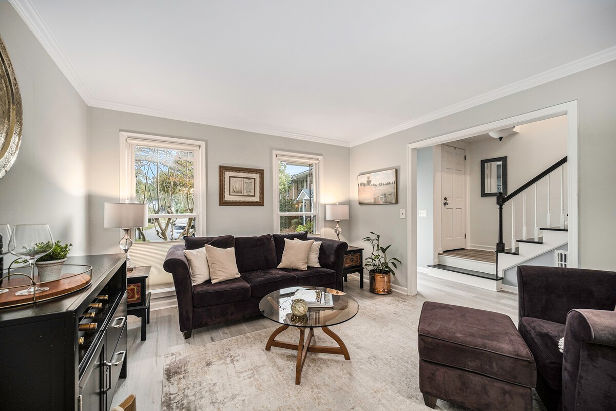 A spacious living area features a dark sectional sofa and coordinating armchair, elegantly arranged around a glass coffee table. Natural light filters through the windows, illuminating a soft rug adorned with neutral tones. A door leads to the entryway, providing a glimpse of the staircase.