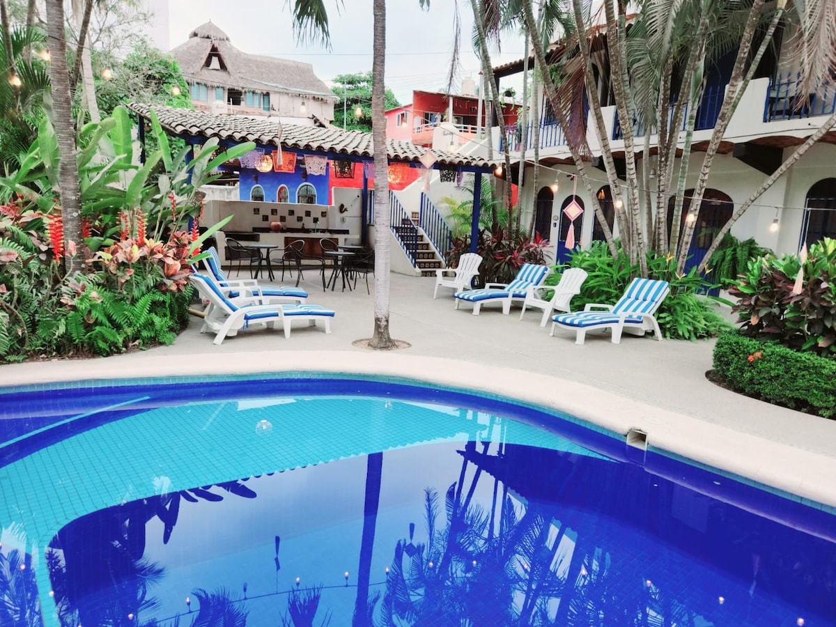 A vibrant pool area features a reflective blue pool surrounded by palm trees and lush tropical plants. Comfortable lounge chairs with striped blue and white designs are placed poolside, creating an inviting space for relaxation. Colorful murals adorn the nearby buildings, enhancing the lively atmosphere.