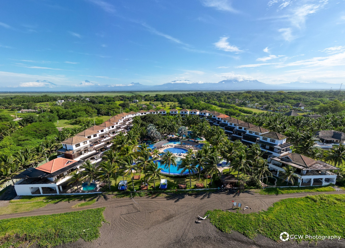 An aerial view of a sprawling resort complex is showcased, featuring multiple buildings arranged around a large central pool. Lush green landscaping, palm trees, and mountain scenery surround the property under a clear blue sky, creating a serene and inviting environment.