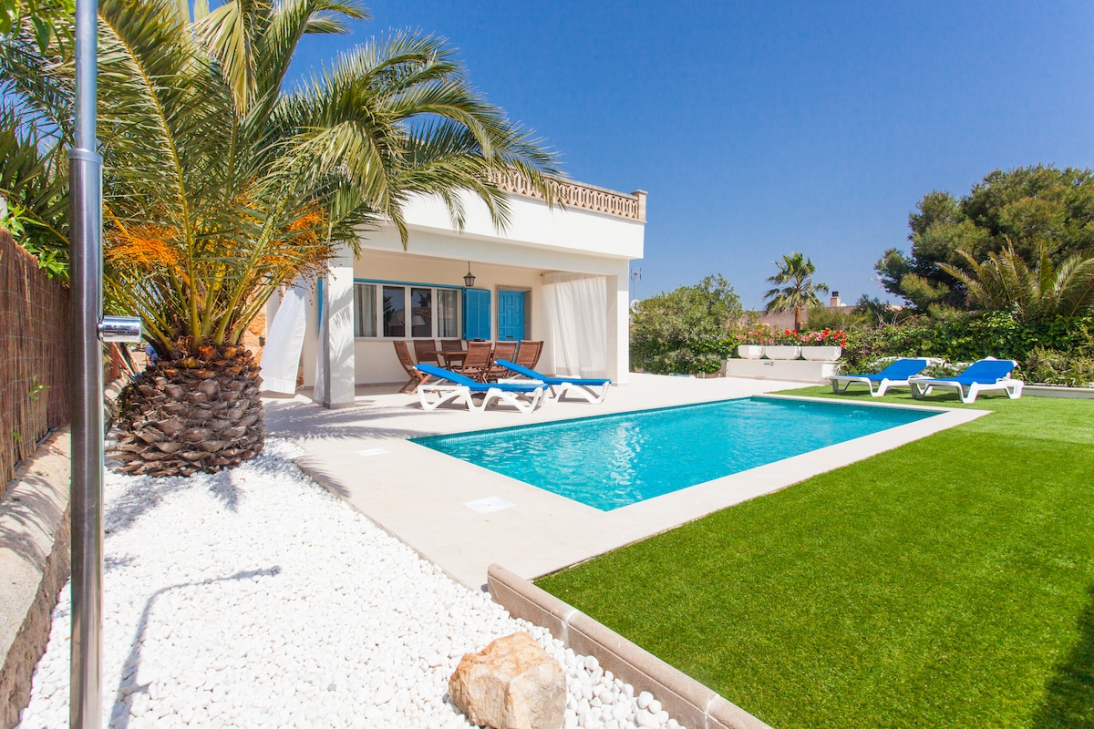 A spacious outdoor area is shown, featuring a bright swimming pool bordered by manicured grass and sun loungers. Tropical palm trees provide shade, while the clean snow-white stones add contrast to the lush greenery. The villa’s exterior is highlighted against a clear blue sky.