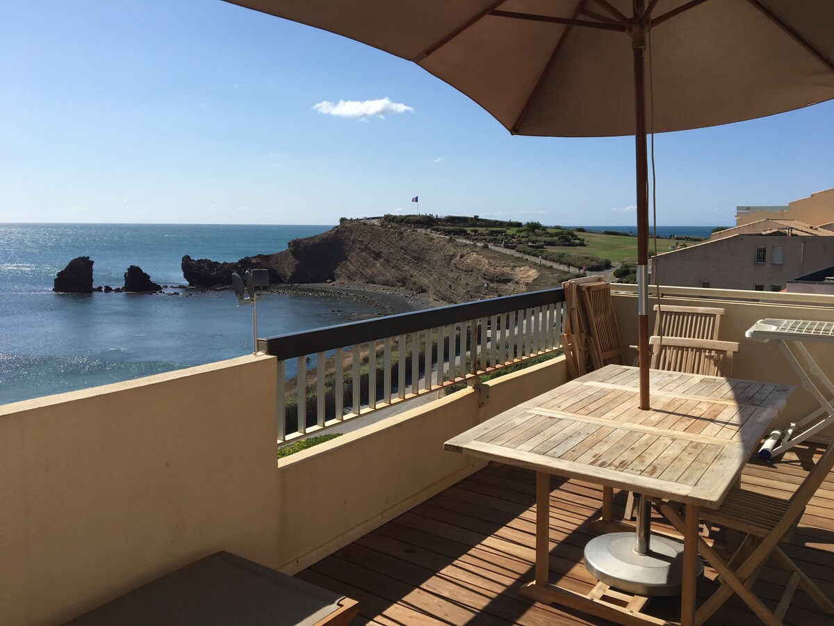 A spacious terrace is shown with a beautiful view of the sea and coastline. A wooden table and chairs are visible, alongside two sun loungers. A large umbrella provides shade, and the serene backdrop of the water and rocky formations adds to the setting.