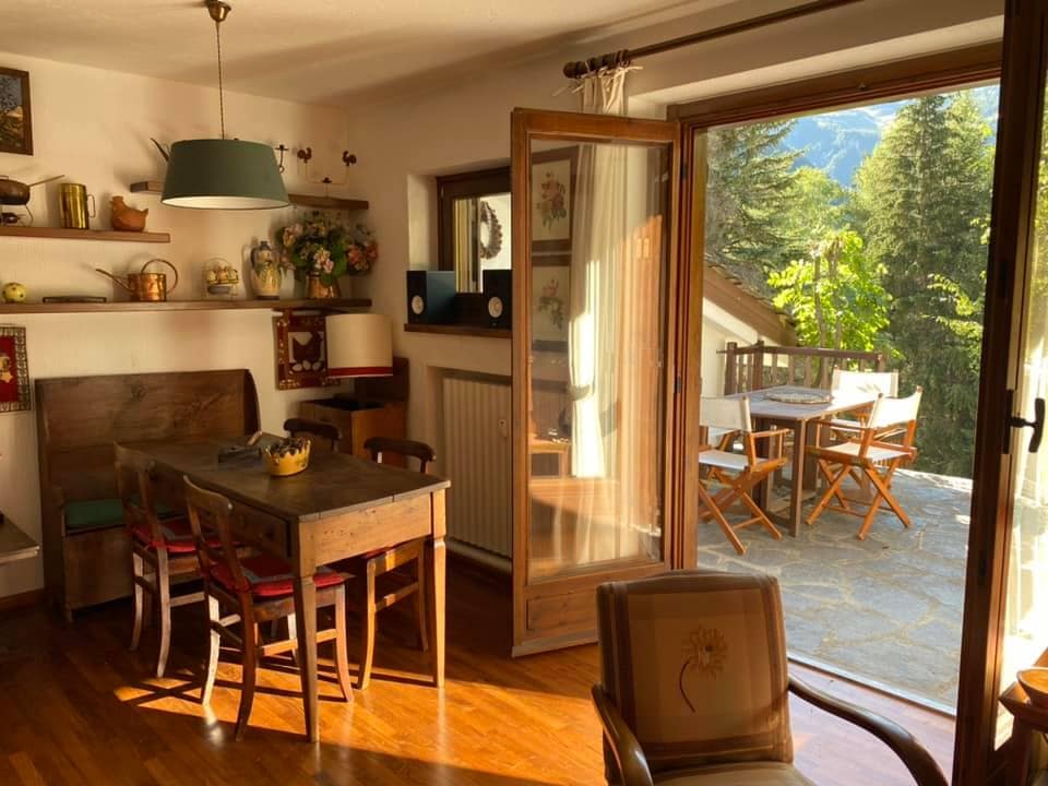 A dining area features a wooden table surrounded by chairs, illuminated by natural light through large glass doors. The space opens to a stone terrace with wooden outdoor furniture, surrounded by trees, providing a peaceful outdoor setting.