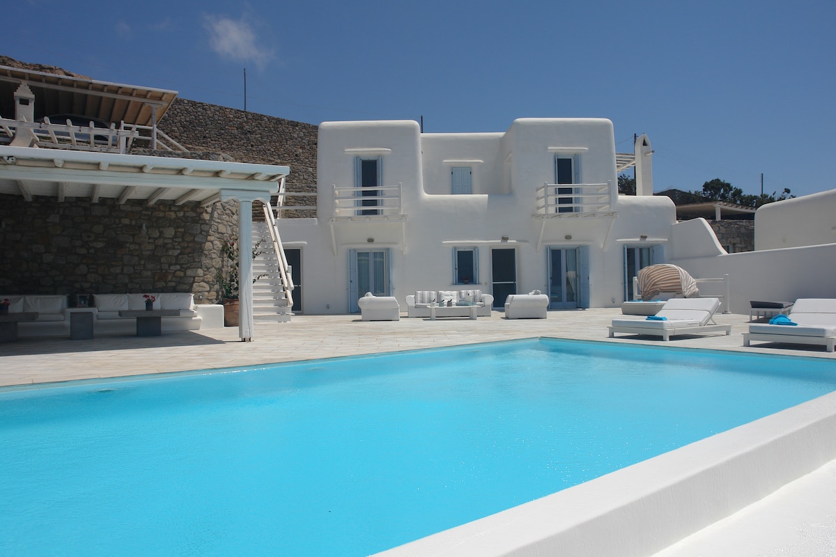 A private pool is framed by a spacious terrace, with sun loungers arranged around it. A two-story white structure with balconies overlooks the area, complementing the clean lines of the architecture. Nearby, a shaded outdoor seating area is furnished with comfortable couches.