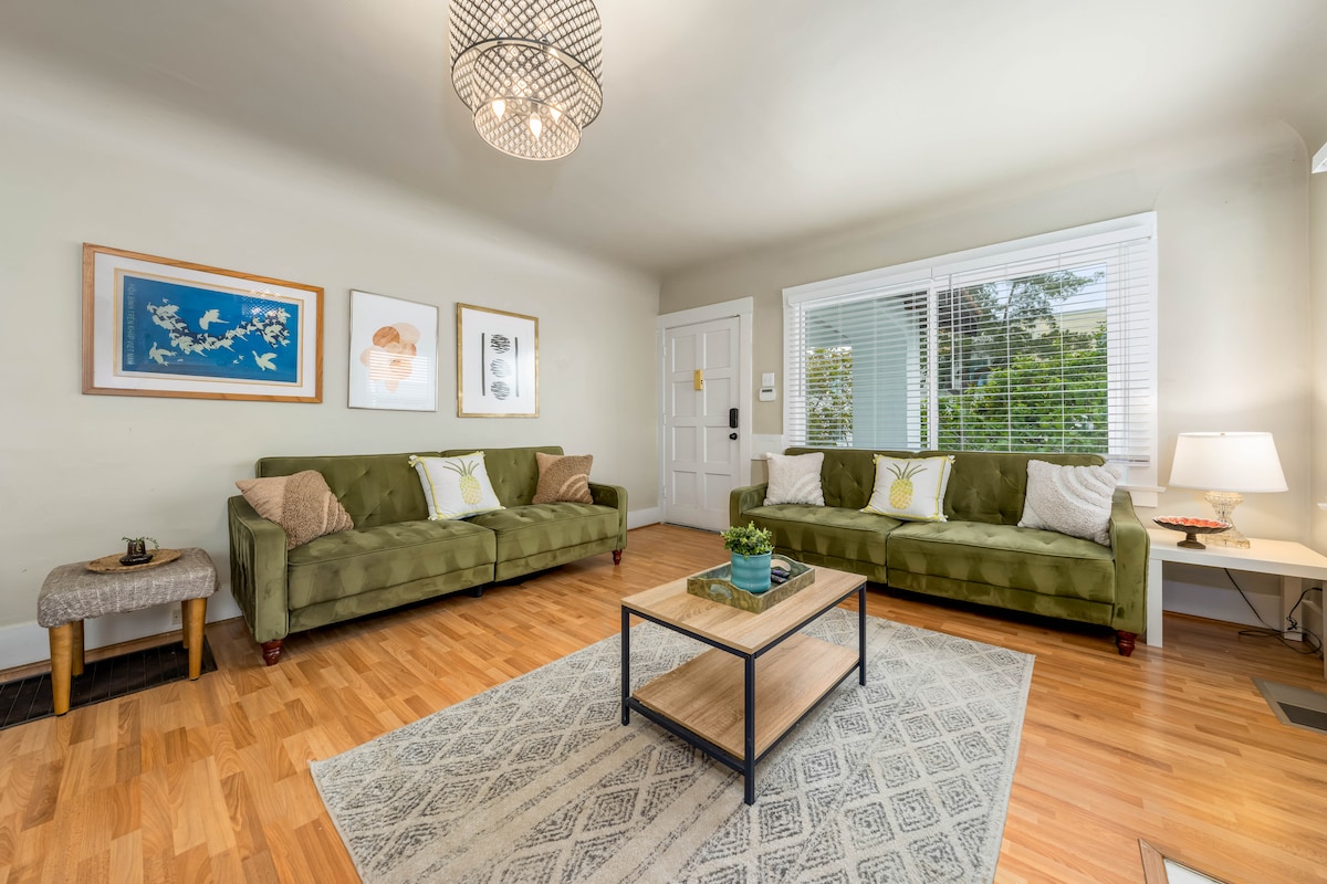 A welcoming living area is highlighted by two green sofas, arranged symmetrically around a central coffee table. Natural light enters through large windows adorned with white blinds, illuminating the hardwood floor. Decorative wall art adds character, while a small plant on the coffee table introduces a touch of greenery.
