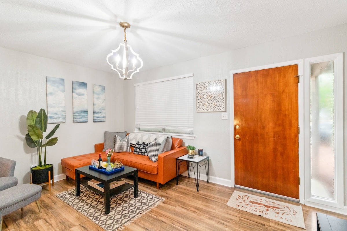 A welcoming living area is presented with a bright, modern light fixture overhead. An orange sectional sofa is positioned beside a small coffee table, which displays decorative items. Three framed artworks adorn the wall, while a large window allows natural light to fill the space.