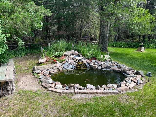 A tranquil pond surrounded by natural stone features is nestled within a lush green yard. Vibrant plants and flowers add color to the landscape, while a rustic wooden bench provides a resting area nearby. Tall trees create a serene backdrop, enhancing the outdoor setting.