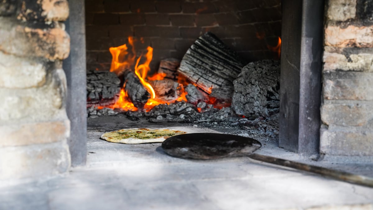 A traditional stone oven is showcased, with flames flickering around hot embers. A flatbread rests on a metal peel at the forefront, while charcoal and ash provide a rustic backdrop. The oven's brick structure adds to the authentic cooking experience.
