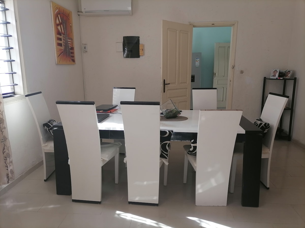 A dining area is depicted, featuring a long rectangular table surrounded by eight high-backed chairs. The table is simple and modern, with a decorative centerpiece. Natural light streams through nearby windows, illuminating the light-colored walls and tiled flooring.