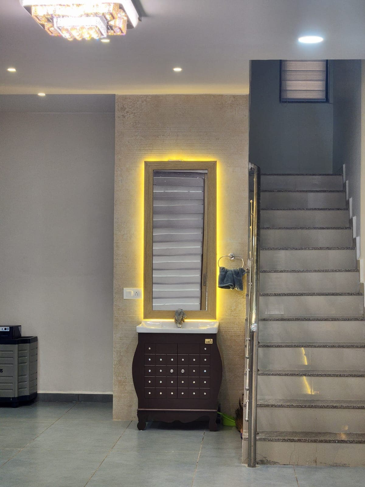 A well-lit entryway features a modern sink with a dark wooden cabinet underneath. A large mirror with backlighting enhances the space, while a staircase with sleek, gray steps leads to the upper level. Soft overhead lighting provides a welcoming ambiance.