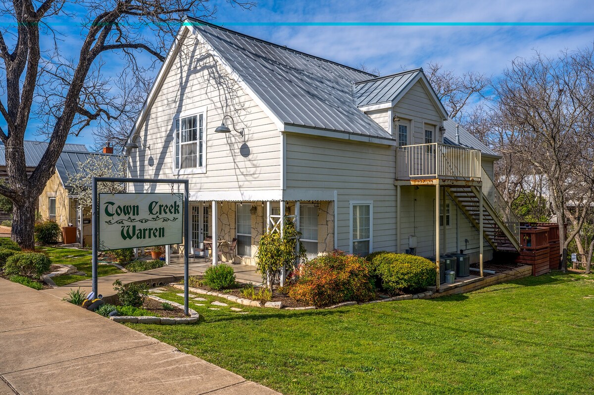 A modern two-story building with a sloped metal roof features light-colored siding and a welcoming sign for Town Creek Warren. The landscaped yard exhibits well-maintained grass and shrubs, with a stone pathway leading to the entrance and a staircase on the right side.